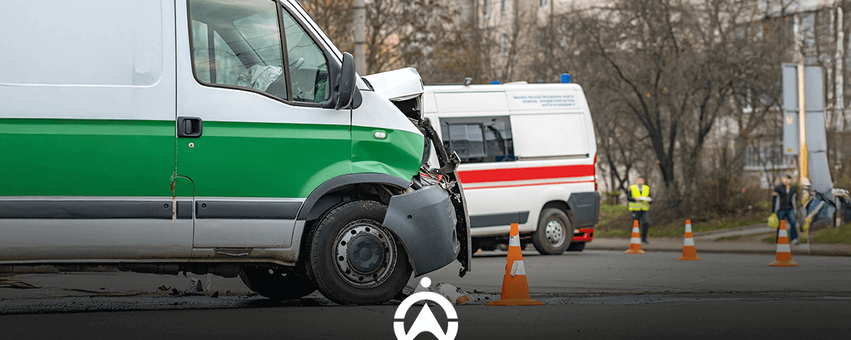 A damaged green and white van in an accident scene, with an ambulance and traffic cones visible in the background.