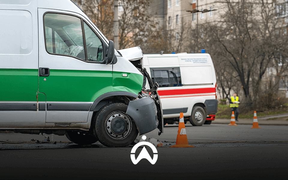 A damaged green and white van in an accident scene, with an ambulance and traffic cones visible in the background.