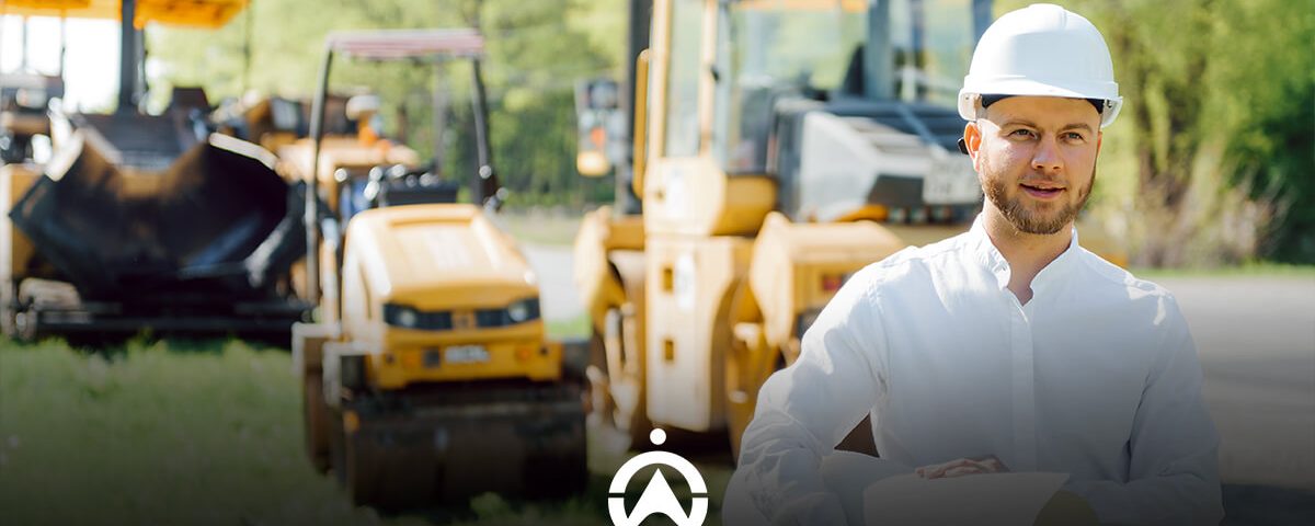 A worker in a white shirt and safety helmet stands in front of yellow construction machinery on a sunny day.