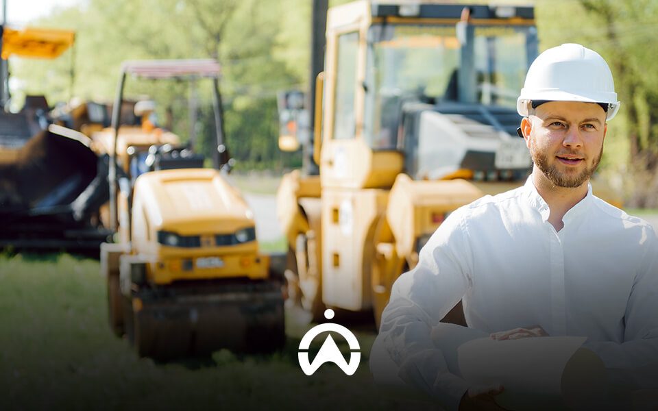 A worker in a white shirt and safety helmet stands in front of yellow construction machinery on a sunny day.