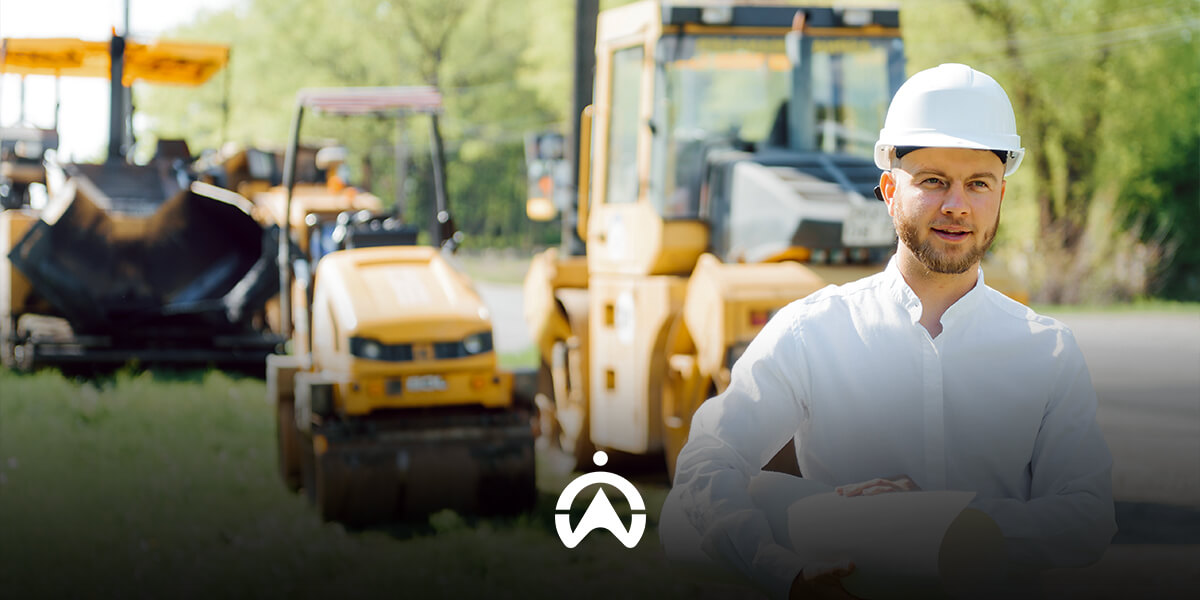 A worker in a white shirt and safety helmet stands in front of yellow construction machinery on a sunny day.