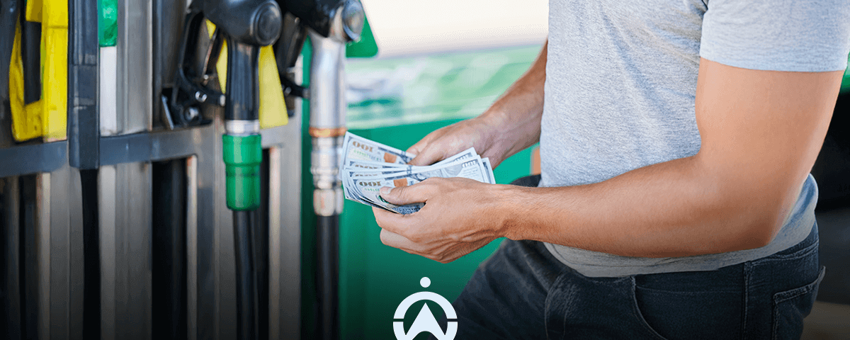 A person holding a handful of cash stands near gas pumps, preparing to pay for fuel.