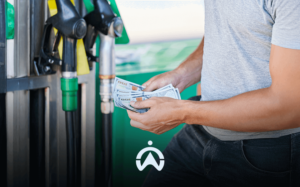 A person holding a handful of cash stands near gas pumps, preparing to pay for fuel.